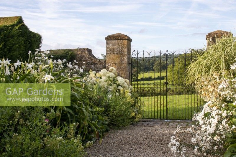 A decorative wrought iron gate between Cotswold stone pillars gives view onto countryside from the White Garden at Bourton House, Gloucestershire.