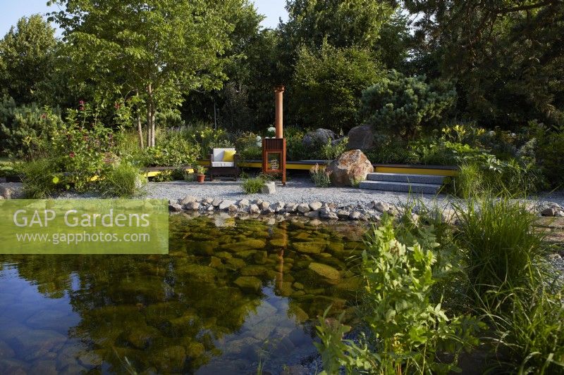 Hurtigruten: The Relation-Ship Garden. Designer: Max Parker-Smith. Hampton Court Garden Festival 2023. Large clear pool with adjacent seating and relaxation area warmed by an outdoor metal log burner. Area of reclaimed fine grey granite setts. Summer.