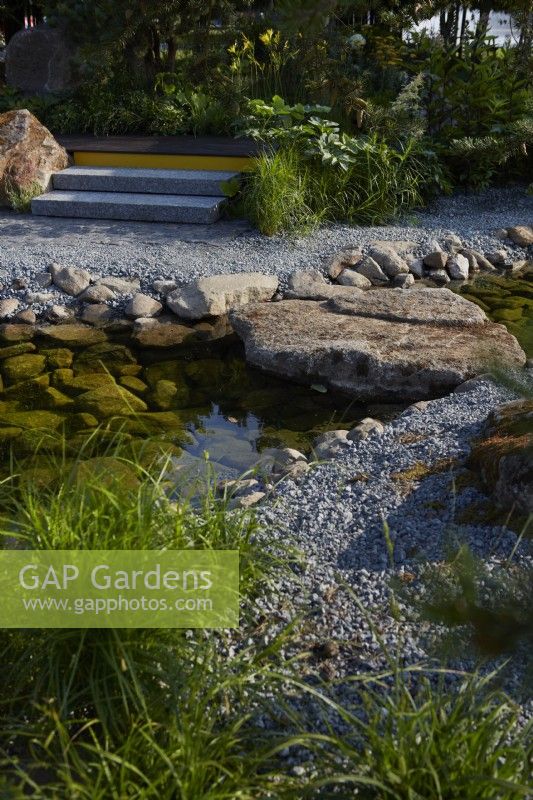 Hurtigruten: The Relation-Ship Garden. Designer: Max Parker-Smith. Hampton Court Garden Festival 2023. Clear pool with large rocks and boulder acting as a bridge. Pathway of reclaimed fine grey granite setts. Summer.