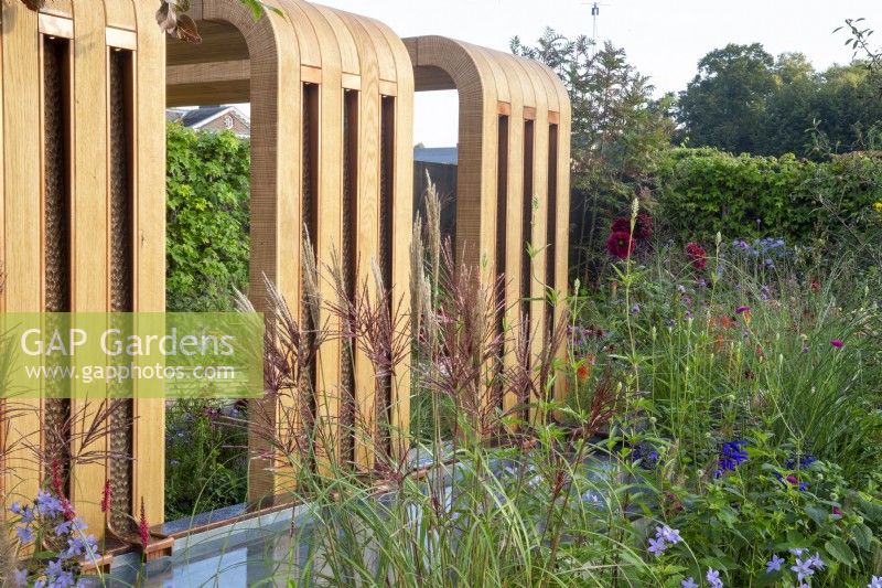 A modern contemporary wooden canopy pergola with water rills in to a pond - planting includes Miscanthus sinensis 'Red Chief'