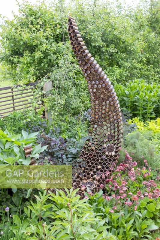 A rusty garden sculpture made from reclaimed, recycled materials with spaces made for an insect hotel - mixed planting of Geum 'Pink Petticoats', Foeniculum vulgare 'Purpureum', Peony, Angelica sylvestris 'Vicars Mead' - syn. 'Vicar's Mead' and a Crataegus monogyna - Common hawthorn hedge