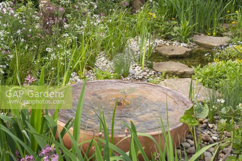 Metal copper bowl water feature in a wildflower garden with pond and stepping stones with marginal planting 