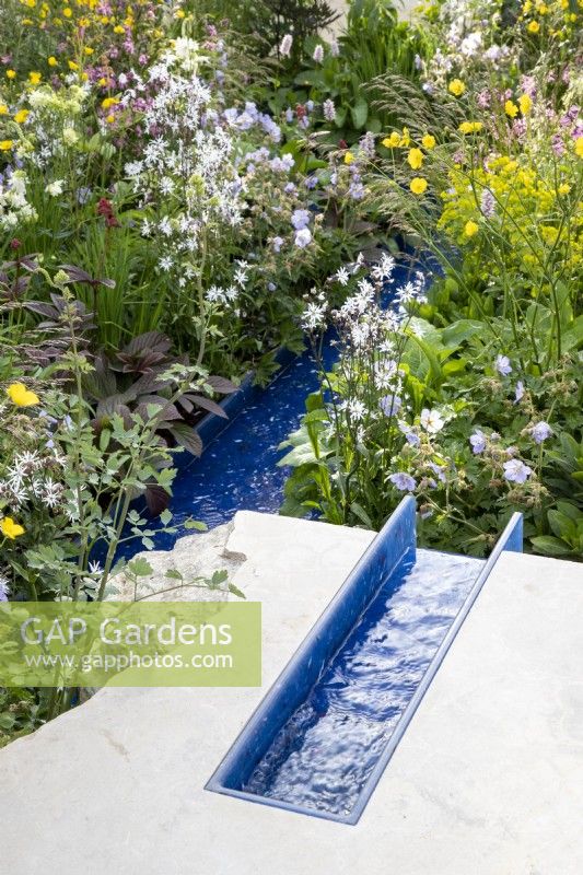 A modern contemporary water rill made from recycled plastics set in stone mixed perennial planting of Geraniums, Lychnis flos-cuculi 'White Robin' and Ranunculus acris 