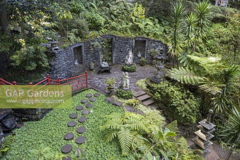 Round stepping stones curve across a pool covered in floating foliage. Steps lead down behind with oriental style red balustrades to a large paved terrace with niches inset with statues. The area is surrounded by tropical planting. Monte Palace Gardens, Madeira. August. Summer