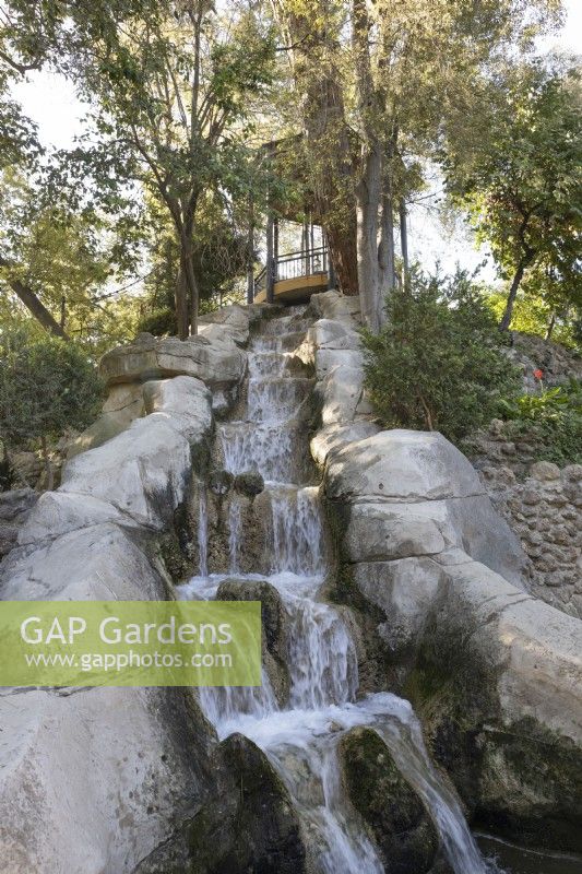 A pergola on top of a mound, with a waterfall tumbling down from under the viewing platform. Various trees and shrubs gorw around it. Parque de Maria Luisa, Seville, Spain. September