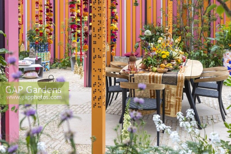 View of the dining area with table and chairs decorated with Indian style ornaments, colourful flowers, fruits and spices. Orange post with sentences in braille on the front. The RHS and Eastern Eye Garden of Unity, Designer: Manoj Malde