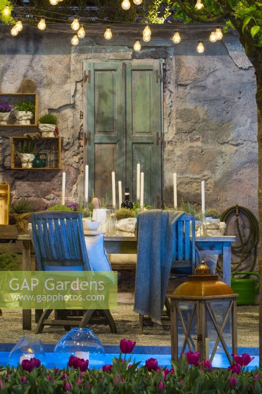 Contemporary Italian courtyard in front of the stone house illuminated by hanging light string of bulbs over decorative table prepared for a spring evening party with candles, glasses and herbs in pot on the gravel surface. Small pond with floating candles. In the foreground, a row of evenly planted purple tulips and lantern. 

