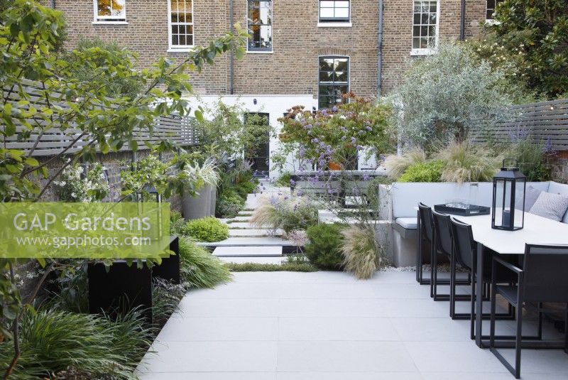 Dining area at the end of the city garden, bordered with Stipa tenuissima, olive tree, Pittosporum tobira 'Nanum' and Verbena bonariensis, with clumps of Hakonechloa macra by the folded metal table