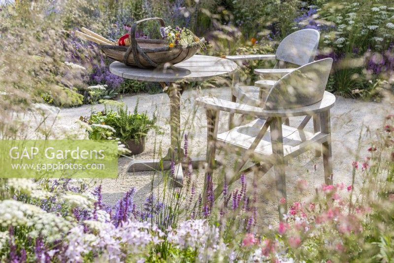 A seating area with a wooden table and two chairs surrounded by a loose meadow style border. RHS Iconic Horticultural Hero Garden, Designer: Carol Klein, RHS Hampton Court Palace Garden Festival 2023