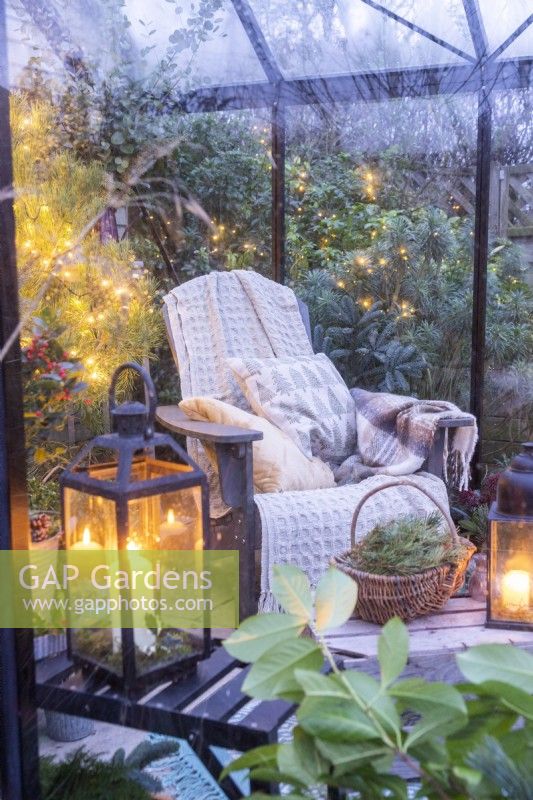 Recycled plastic chair with blankets and cushions, wooden crate and plants adorned with fairy lights inside a greenhouse