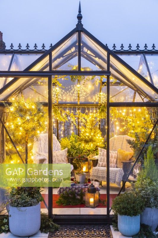Large greenhouse furnished and decorated with recycled plastic chairs, fairy lights and mixed planting on patio