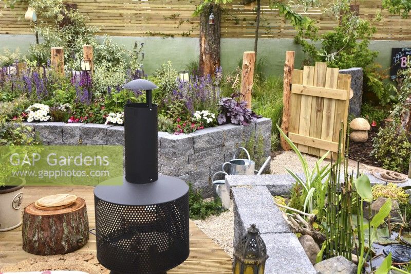 View picnic area on wooden deck with modern chiminea and mini pond to a raised bed made of  Connemara walling system in a woodland inspired garden. Planted with  Leucanthemum superbum, Salvia Nemorosa and aquatic plants. June
