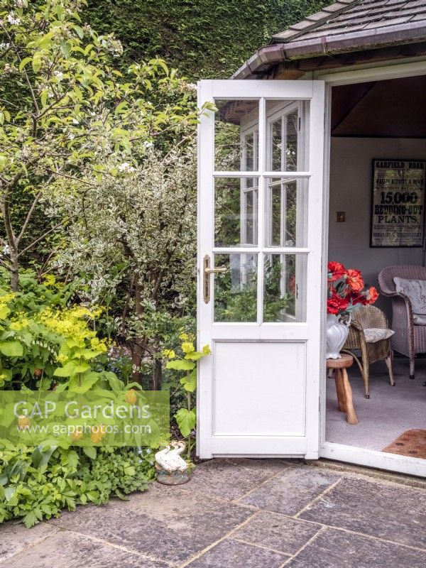 Summerhouse doorway next to a mixed border featuring Smyrnium perfoliatum, Rubus deliciosus, Euonymus fortunei, Myosotis, Polygontatum and Tulipa 'Brown Sugar'