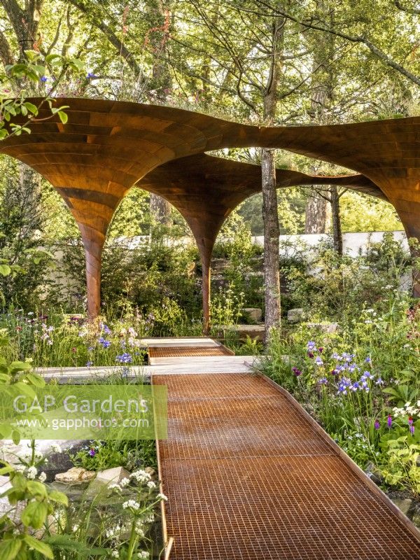 Open metal decking bridges lead to a large structural pavilion alongside perennial and aquatic planting. WaterAid Garden, Designers: Tom Massey  and  Je Ahn, Gold medal, RHS Chelsea Flower Show 2024, Sponsor: Project Giving Back
