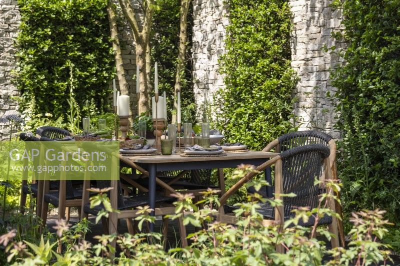 An outdoor dining table sheltered by a stone wall and shrubs - Killik  and  Co: Money Doesn't Grow on Trees Garden - designer Baz Grainger - RHS Chelsea Flower Show 2024