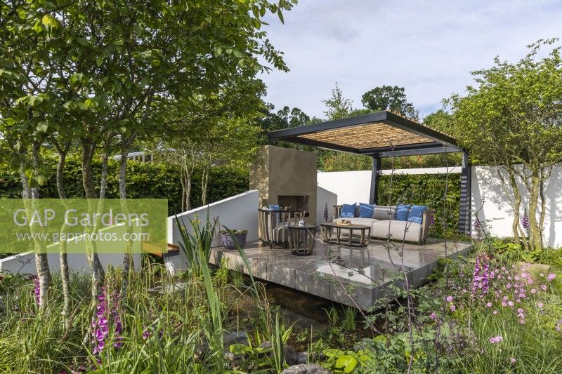 A modern garden with pond, lounge patio with fireplace and comfortable outdoor furniture enclosed within planting and cocooned by a contemporary openwork steel pergola. Designer: Joe Eustace 