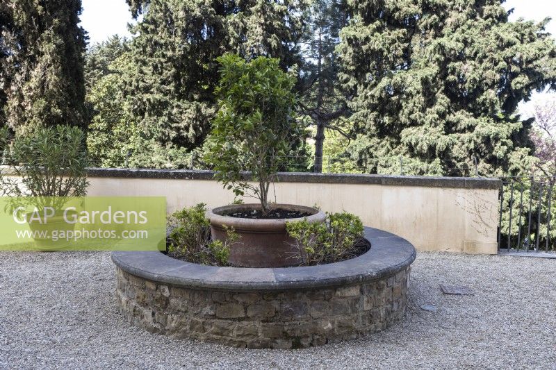A stone bench encircles a large terracotta planter on a gravelled terrace. Bardini Gardens. Florence. Spring, April. 