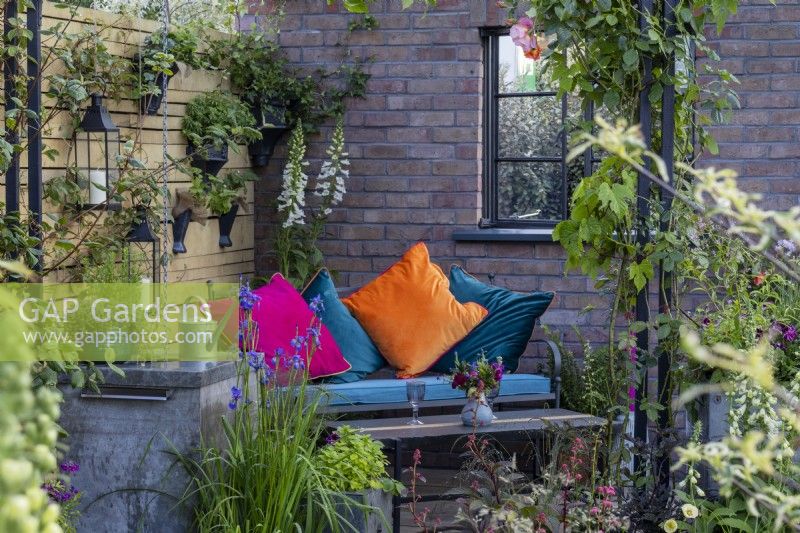 An outdoor seating area is set between a brick wall and fence made from salvaged timber with wall containers of plants - 'Flood Re: The Flood Resilient Garden' - designers Naomi Slade and Ed Barsley - RHS Chelsea Flower Show 2024