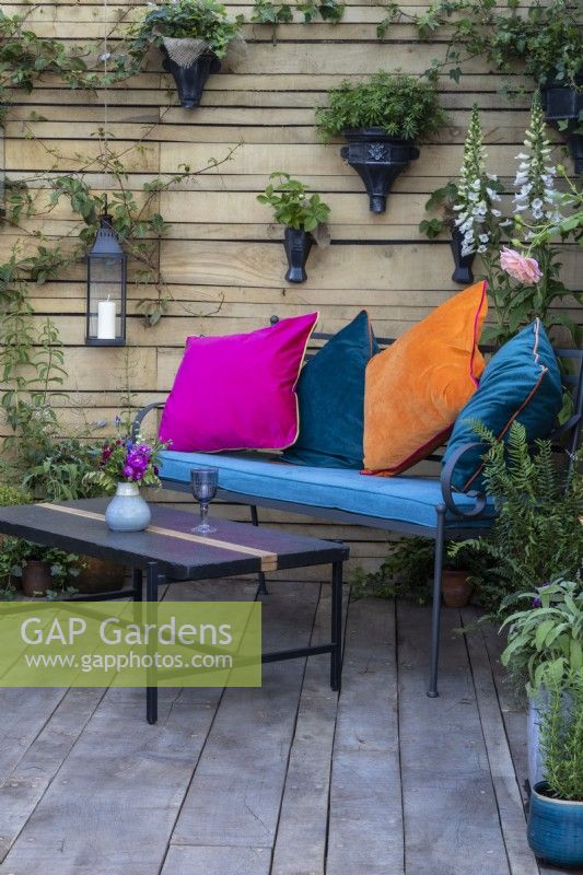 A metal bench and table with colourful cushions on a wooden deck with a salvaged timber fence behind and wall containers of plants - 'Flood Re: The Flood Resilient Garden' - designers Naomi Slade and Ed Barsley - RHS Chelsea Flower Show 2024