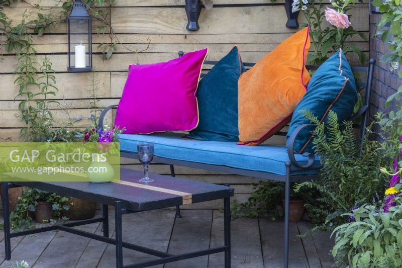 A metal bench and table with colourful cushions on a wooden deck with a salvaged timber fence behind and containers of plants around - 'Flood Re: The Flood Resilient Garden' - designers Naomi Slade and Ed Barsley - RHS Chelsea Flower Show 2024