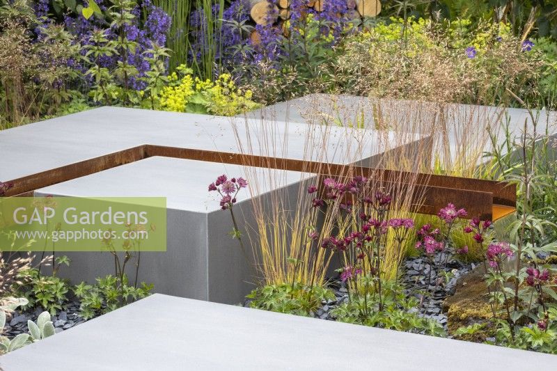 Geometric porcelain blocks are set against perennial planting including Deschampsia cespitosa 'Goldtau' and Astrantia 'Moulin Rouge' with a slate chipping mulch A Corten steel rill ends with a fall of water - Eco Oasis Garden - designer Dan Hartley - BBC Gardeners' World Live 2024