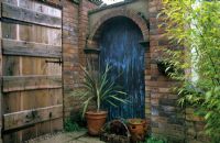 Corner of patio with copper clad alcove and rustic oak door. Cordyline in container. 