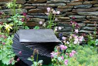 Decorative slate spiral feature and stone wall with Astrantia major 'Hadspen Blood', Sanguisorba and Aquilegia vulgaris.