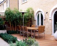 Wooden terrace beside house with table and chairs, Phyllostachys vivax 'Aureocallis' and Buxus