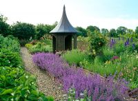 Willow summerhouse with lead covered roof in cottage garden with willow fences and raised beds, Nepeta 'Walker's Low', Delphiums, roses, Persicaria bistorta 'Superba' and Alliums