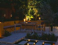 Aluminium table and chairs on patio surrounded by yellow rendered walls with raised beds and rill. Evening lighting with candles and lanterns. 