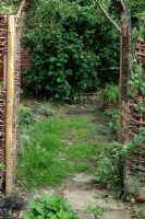 Neglected path in need of repair viewed through willow archway with Corylus avellana 'Contorta' in background