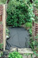 Laying down weed suppressant fabric over neglected path in need of repair, viewed through willow archway. Corylus avellana 'Contorta' in background.