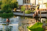 Couple enjoying swimming pond on warm summer's evening with decking and seating in background - Bressingham, Norfolk.