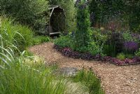 Herbaceous border and ornamental grass border with bark chippings path leading to a wooden arbour with swinging wooden seat - The Rainbow garden, 'Wedgwood', Hesketh Bank, Lancashire