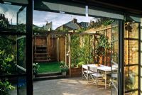 Small urban courtyard garden with pergola, lawn, childs play area and dining area viewed from inside house - Acton, London

  