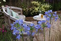 Curved wooden seat with small table on stone patio surrounded by light colourful planting including blue Agapanthus in the 'A Tranquil Retreat' garden, Exhibitor - Garden Creations, Sponsor - Acorn Stone Merchants, Tatton Flower Show 2008