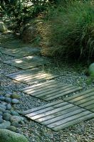 Wooden panels inset in to a gravel and pebble path in a grasses and bamboo garden. Designed by Alan Titchmarsh at Barleywood, Hampshire.