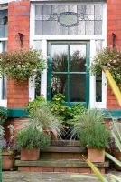 Green back door to a traditional Victorian red brick house with stained glass surround. Raised steps with pots of geranium, grasses and herbs