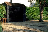 Concrete garage painted black to imitate black timber in urban front garden with gravel and timber driveway. Existing Cherry tree and Laurel hedge.
