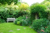 Lawn with stepping stones leading to gate in garden wall, herbaceous border with roses and jasmine and white garden bench - New Square, Cambridge