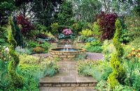 Formal water garden with raised circular pond and fountain on patio on sloping site with mixed borders and sprial topiary