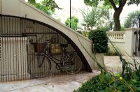 Corten steel to the side of the stairs creates a weather cover for a bicycle and  conceals the utility meters, ivy grows over the entrance pillars 