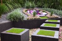 Raised, turf covered slate stepping stones lead through a sunken bed of Impatiens 'Cupido Pink' towards a seating area surrounded by sharp and spiky planting of Cordyline australis, Agave americana, Yucca gloriosa, Yucca recuvifolia, Yucca floribunda, Chamaerops humilis cerifera and Astelia chathamica 'Silver Spear' in the The Quilted Velvet Garden, sponsored by Quilted Velvet and Hortus Infinitus - Silver Flora medal winner at RHS Chelsea Flower Show 2009