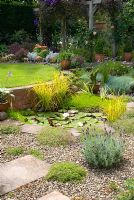 View of secluded back garden with raised lawn, pond planted with Nymphaea - water lily, Iris pallida 'Variegata', Carex elata 'Aurea' and Myriophyllum, with adjacent 'beach area' with Thymus serpyllum 'Coccineus', Lavender 'Hidcote', Dianthus and Osteospermum, and beyond, pergola with Clematis jackmanii 'Superba', Rosa 'Compassion' and purple foliage bed with Cotinus coggygria 'Royal Purple', Berberis thunbergii 'Atropurpurea Nana', Cineraria maritima, Alstroemeria 'Devotion' and Campanula persicifolia alba at Gorse Way, NGS garden, Lancashire