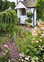 Borders of herbaceous perennials black and white barn door with well planted hanging basket with flowers in profusion, packed into an idyllic English cottage garden, at Grafton Cottage, NGS , Barton-under-Needwood Staffordshire