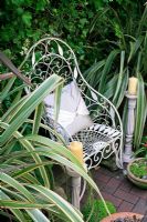 Ornate white wirework chair flanked by variegated Phormiums and candlesticks makes a striking focal point in a secluded town garden