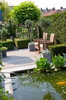 Small formal urban garden with raised pond and water spouts. Patio beyond with Olea europaea - Olive trees in pots. Clipped Buxus - Box hedges. Catalpa bignonioides 'Nana' tree in background. Taxus- Yew hedge. 

