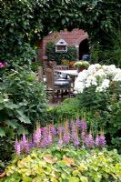 Mixed border of Astilbe, Phlox paniculata 'Fujiyama' and Epimedium with view to seating area and house through ivy covered archway