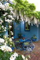 Mosaic tiled patio in small cottage garden with climbing Rosa and Wisteria on pergola. Blue painted house and furniture. No. 11, Christchurch, New Zealand