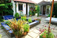 Paved courtyard with raised pool and seat clad in blue glazed tiles. Phormium in container.